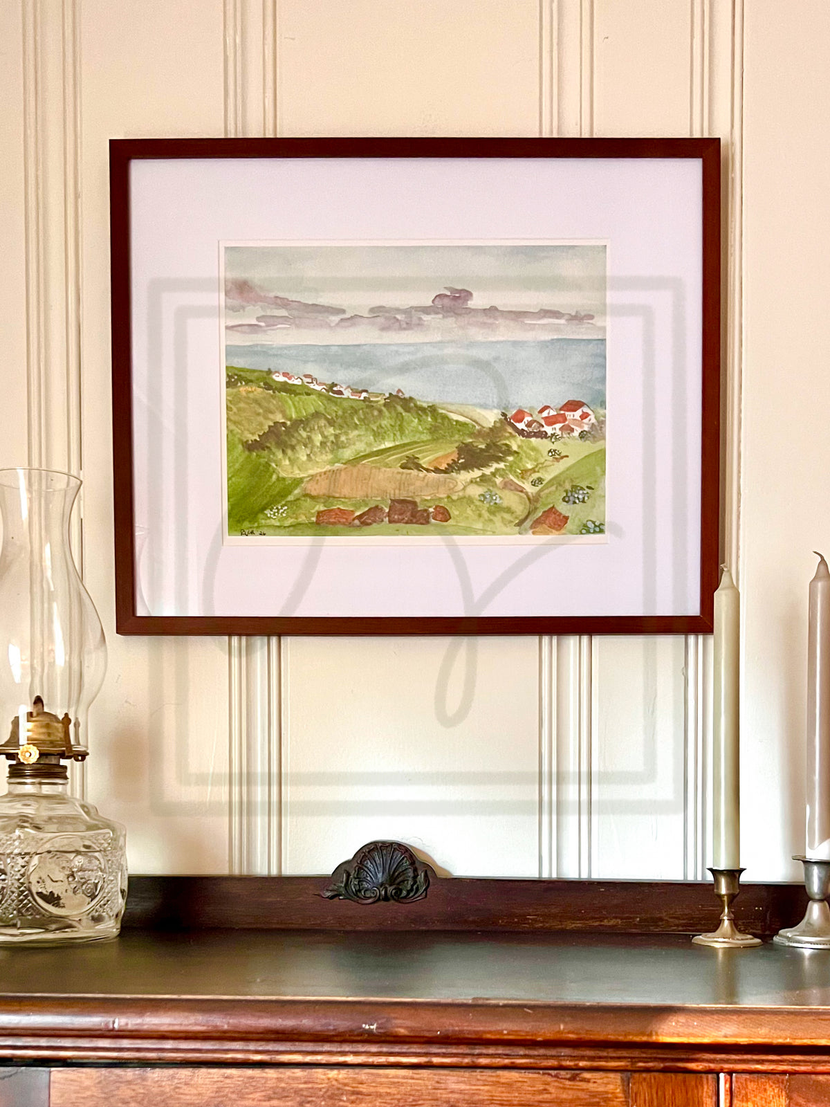 A framed watercolor landscape print of a coastal village with green hills, red-roofed houses, and a blue sea, displayed on a wooden console against a cream paneled wall.